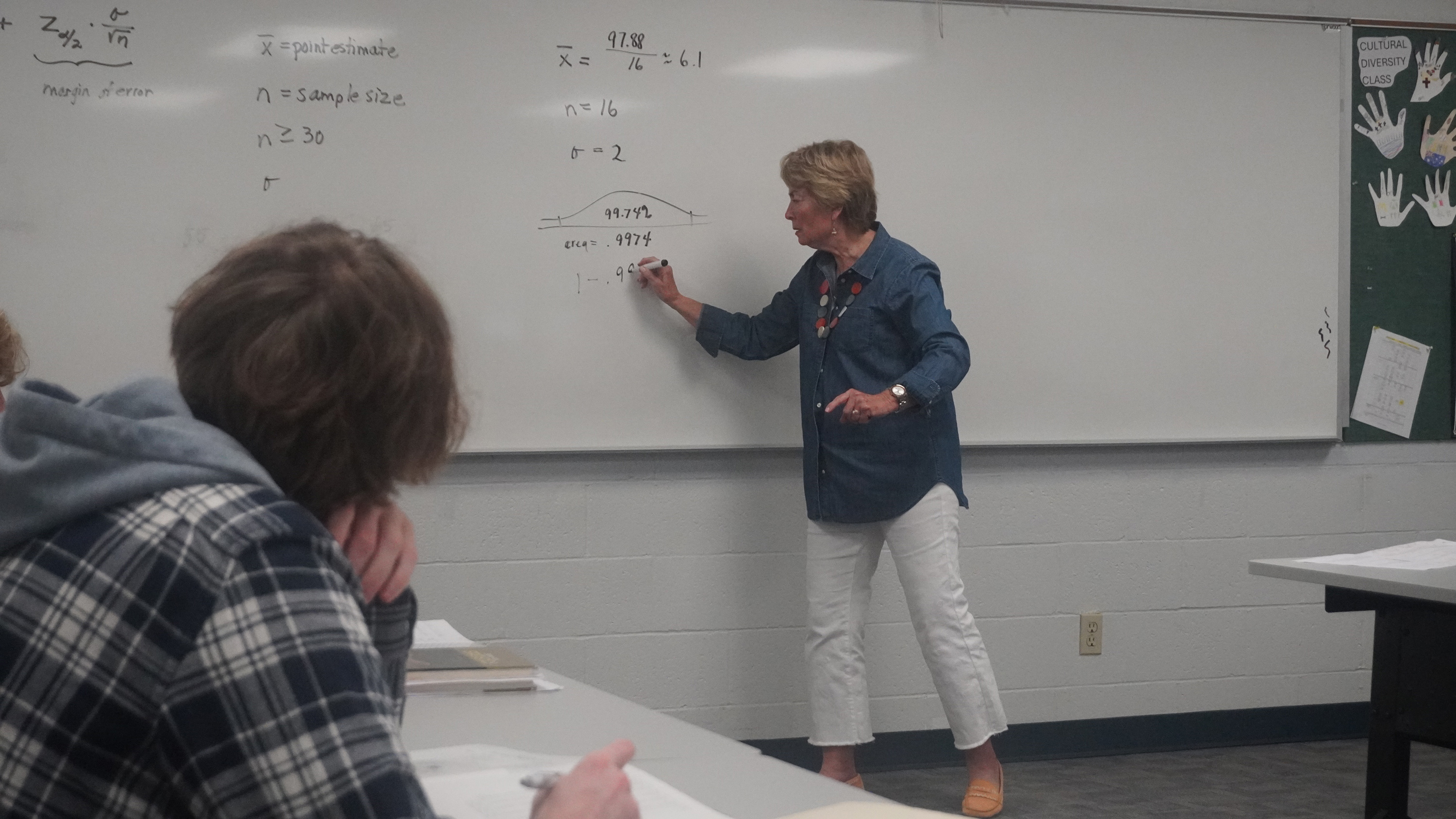 Linda Monge teaches a math lesson, writing equations on a whiteboard while students listen attentively. The classroom setting emphasizes hands-on learning and engagement, with mathematical concepts and problem-solving on display.