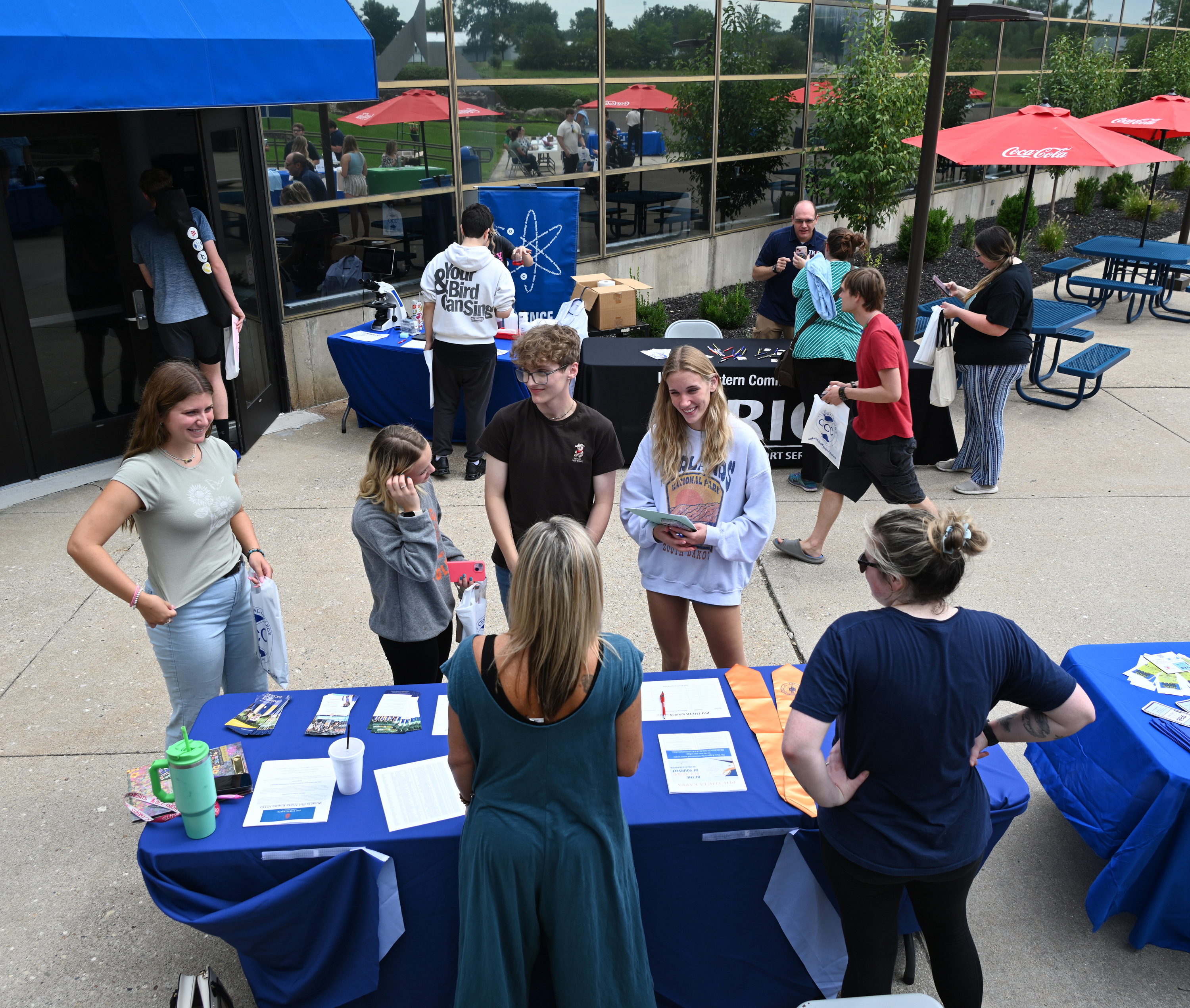 Students and staff gather outside at an informational event, engaging with representatives at tables covered in brochures and materials. Attendees converse, explore opportunities, and enjoy the vibrant outdoor setting with blue and red umbrellas. The event fosters community engagement and student involvement.