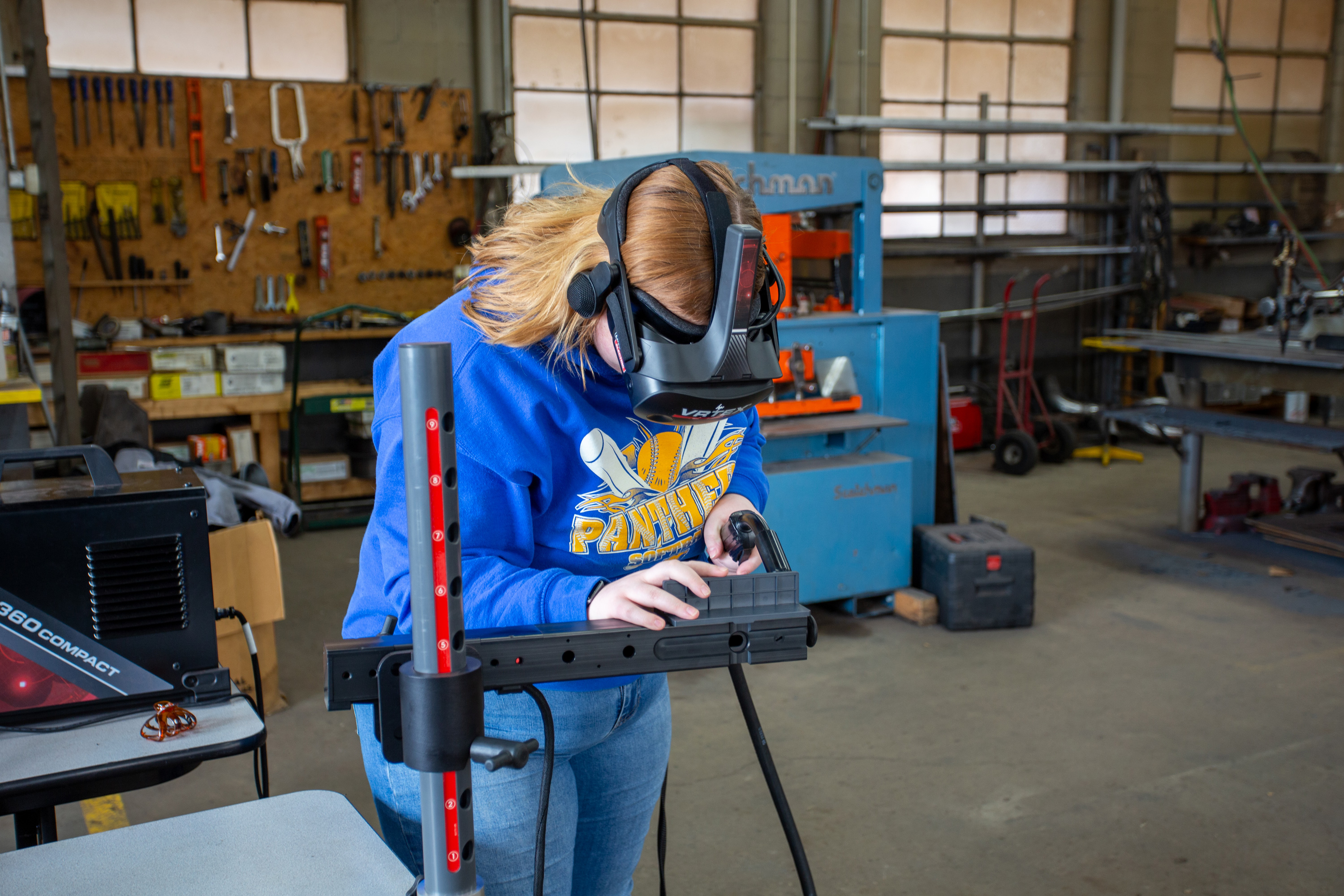 A student wearing a virtual reality welding headset practices welding techniques in a workshop. The VR system provides a hands-on learning experience in a safe environment, surrounded by tools and equipment for technical training.