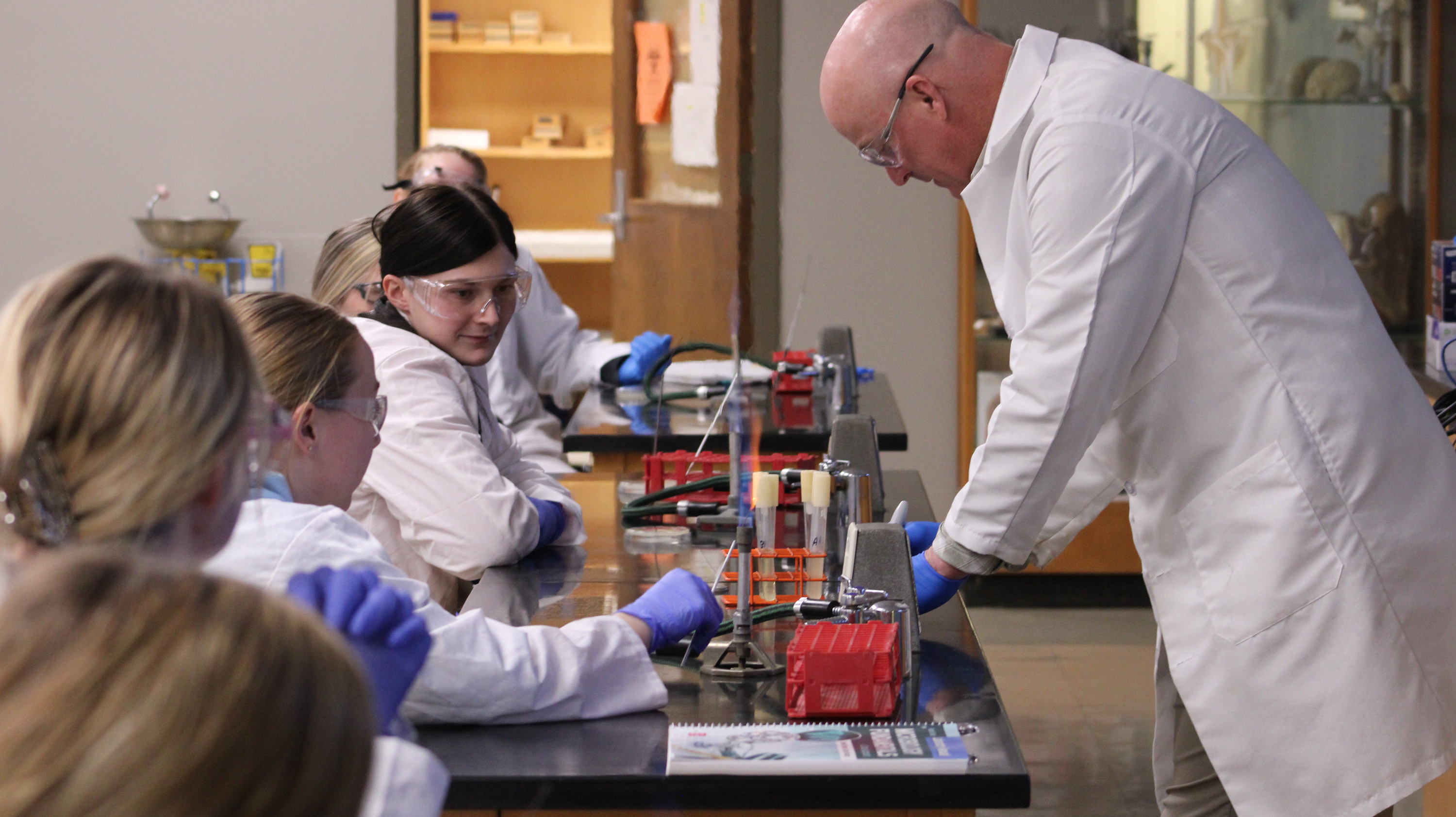 Dr. Todd Gill, wearing a white lab coat, demonstrates an experiment with a Bunsen burner as students, also in lab coats and safety goggles, observe closely. The hands-on lab experience fosters engagement and learning in a collaborative classroom setting.