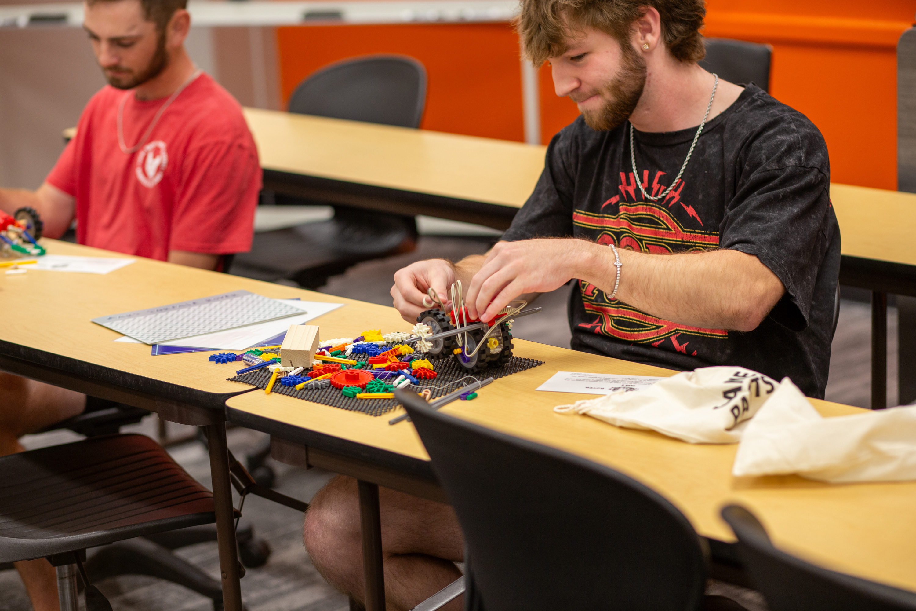 Two students in a classroom engage in a hands-on activity using colorful building materials and mechanical components. One student, wearing a black graphic t-shirt, assembles a project with focus, while another in a red shirt works in the background. The classroom setting encourages creativity and problem-solving.