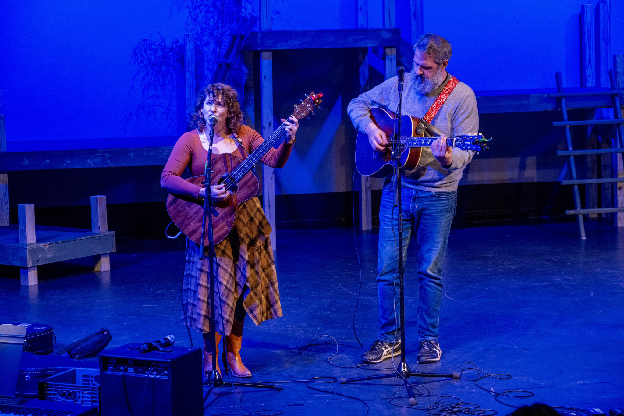 Two musicians perform live on stage under blue lighting, each playing an acoustic guitar. The singer, dressed in a plaid skirt and boots, engages the audience, while the other musician accompanies with intricate guitar work. The set design creates an intimate and artistic atmosphere.