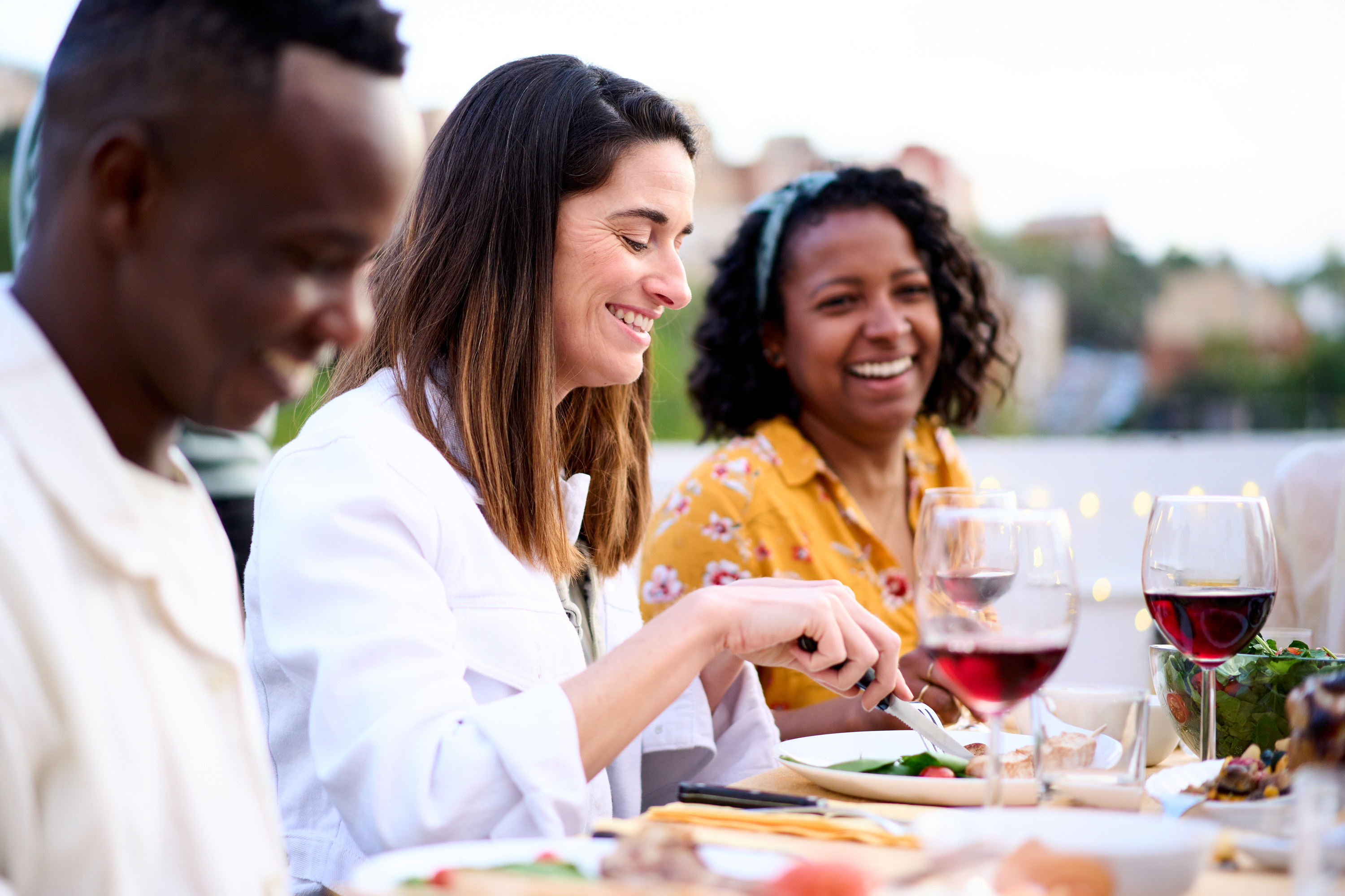 A group of friends enjoys an outdoor meal together, smiling and engaged in conversation. The focus is on a woman in a white jacket, cutting her food while laughing with those around her. The warm lighting and blurred background create a relaxed and inviting atmosphere.