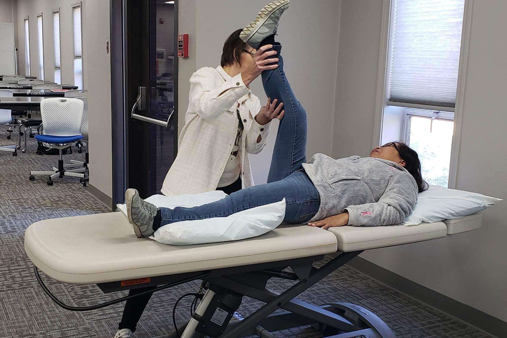 A physical therapy session in a classroom setting, where an instructor demonstrates a leg stretch on a patient lying on an adjustable exam table. The hands-on learning environment provides students with practical experience in rehabilitation techniques.