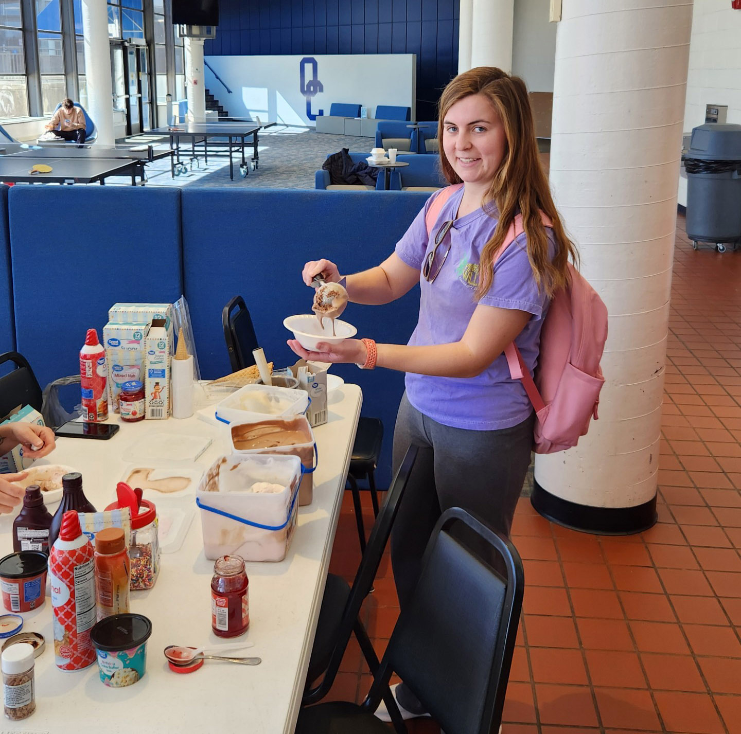 A student in a purple shirt and pink backpack smiles while scooping ice cream at a toppings bar. The table is filled with ice cream, syrups, and sprinkles, creating a fun and welcoming atmosphere. The background features a student lounge with seating, a ping-pong table, and a relaxed social setting.