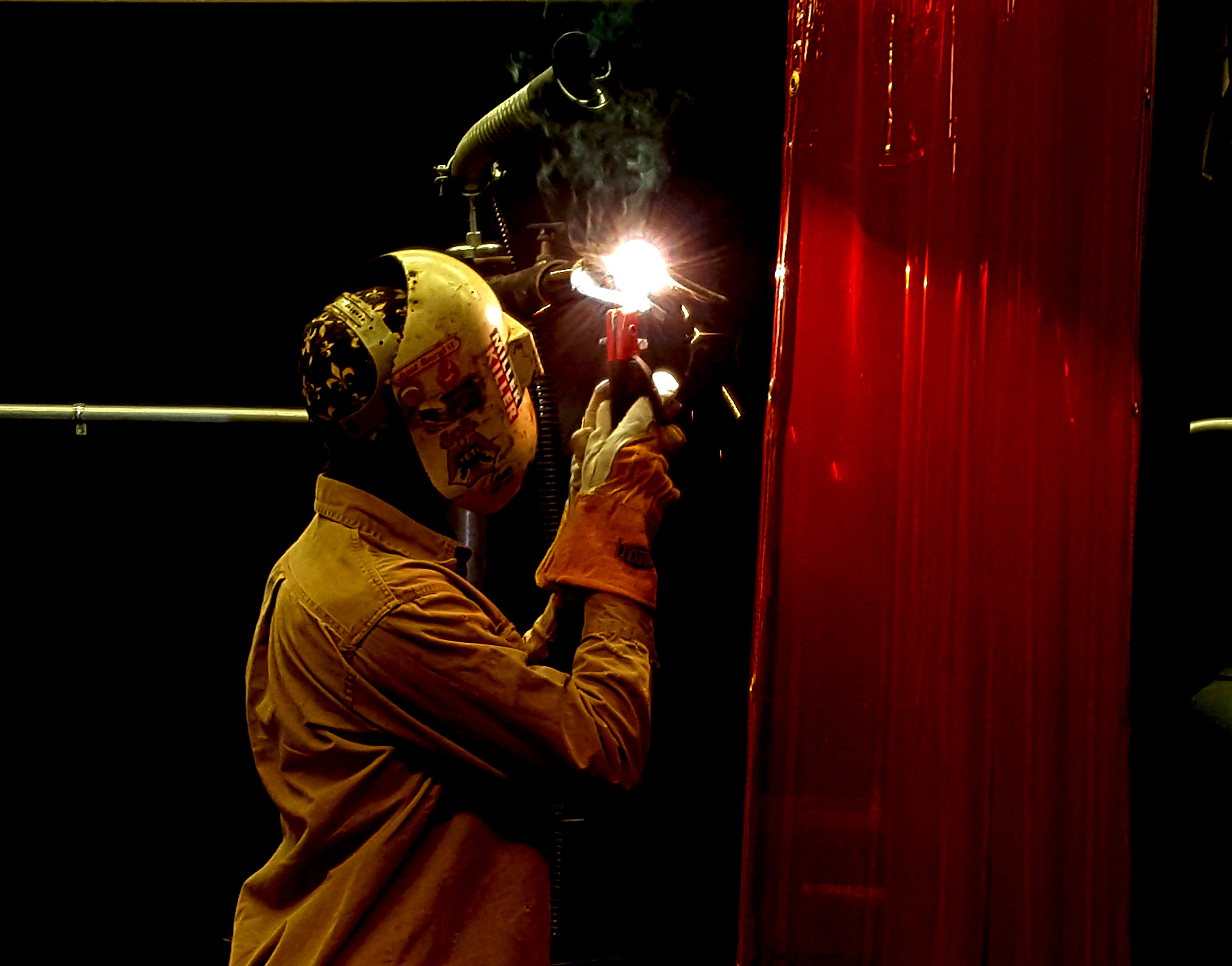 A welder wearing protective gear, including a helmet and gloves, fuses metal pieces in a dimly lit workshop. Bright sparks and smoke rise from the welding torch, illuminating the work area. A red protective curtain is partially visible, providing safety in the workspace.