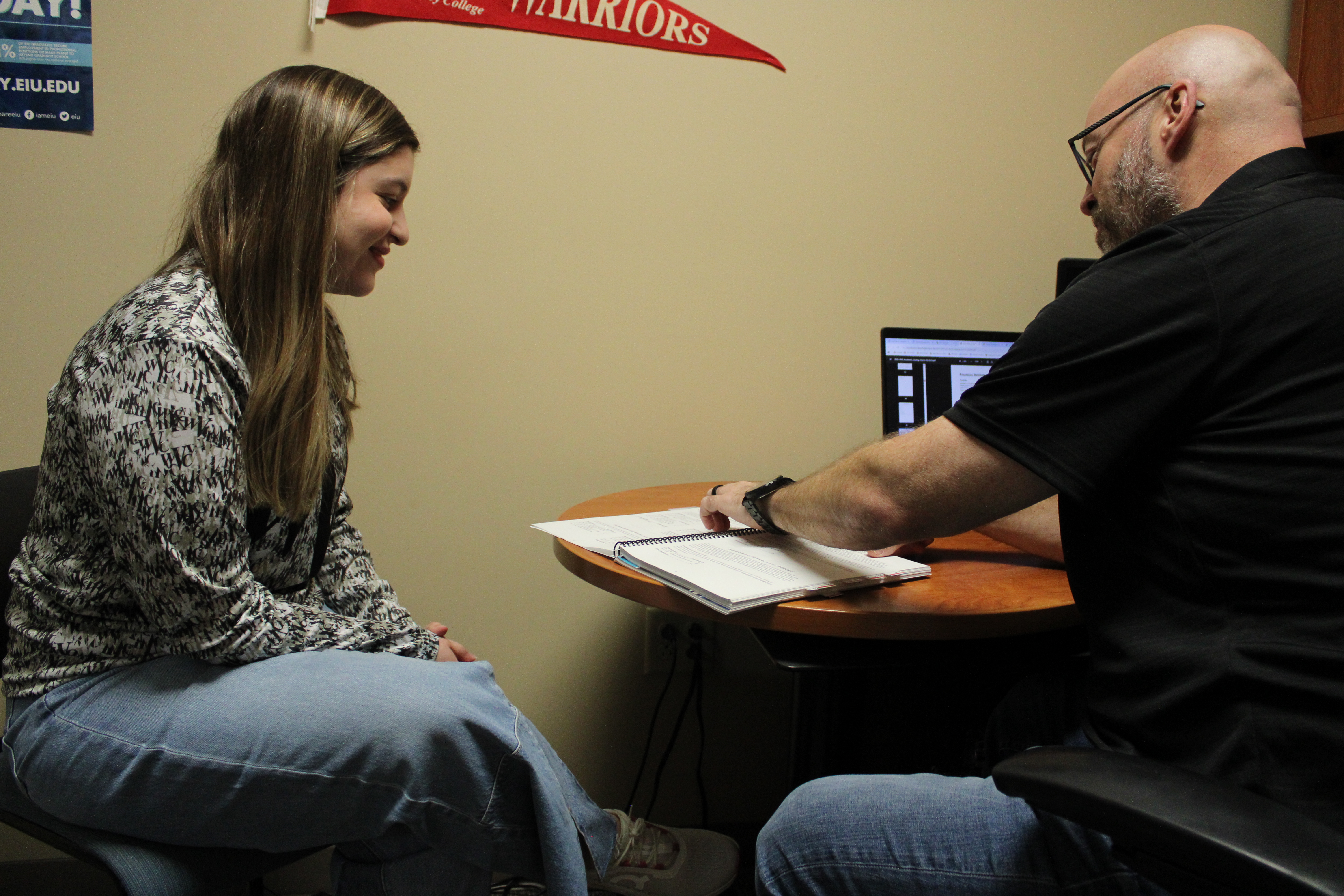 Two individuals seated at a small round table in an office setting, reviewing a spiral-bound notebook. A computer monitor displaying a dark interface is visible on the desk, along with some papers. The wall behind them has a red pennant with the word ‘Warriors’ and part of a blue poster.
