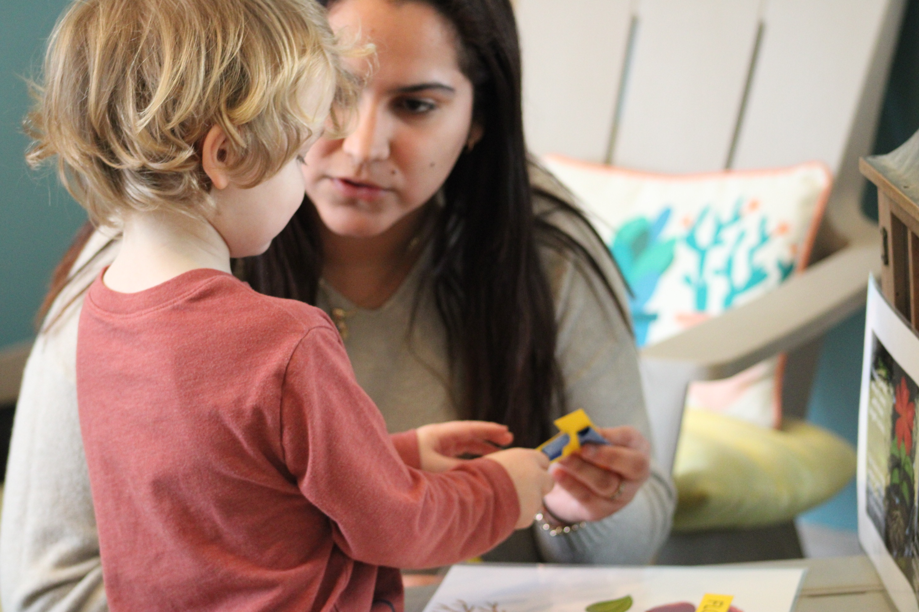 A young child stands at a table holding small colorful blocks while a WVC student seated nearby guides them. The table has papers with pictures, and a cushioned chair with a patterned pillow sits in the background.