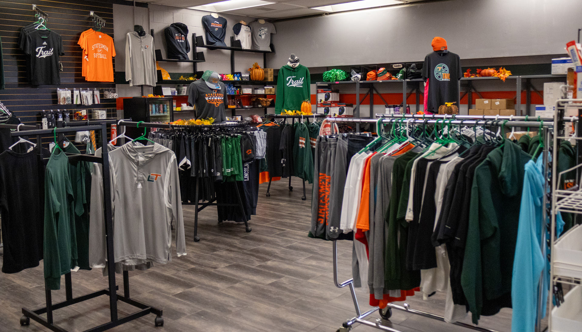 Interior of the Statesmen Store at Lincoln Trail College, featuring racks and displays of college-branded apparel in green, orange, black and gray, with autumn decorations throughout the store.
