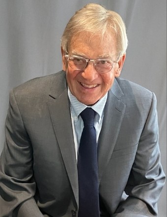 Portrait of Bill Thomas smiling while wearing glasses, a gray suit, light blue shirt and dark tie against a neutral gray background.