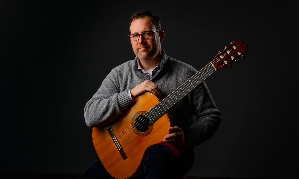 A portrait of Ben Gateno seated holding an acoustic guitar