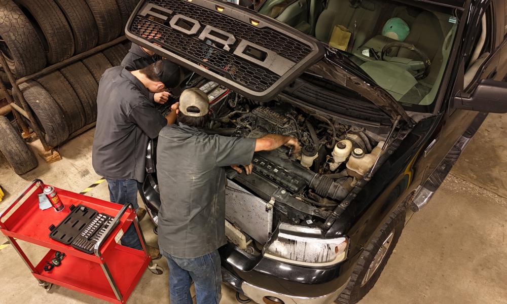 Two automotive students work under the hood of a black Ford truck in a workshop, focusing on diagnosing or repairing the engine. A red utility cart nearby holds tools and lubricant, while a stack of tires adds to the workshop setting. The students, wearing black shirts and caps, demonstrate teamwork and hands-on training, emphasizing the practical skills developed in an automotive education environment.