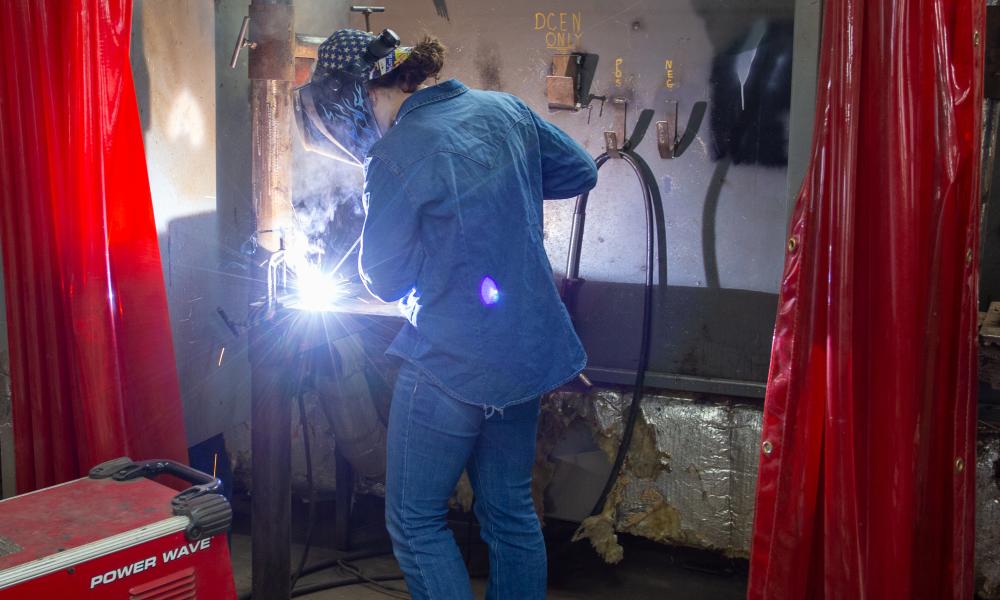 A welding student wearing a protective helmet and denim clothing is actively welding a metal piece in a workshop. Bright sparks and a welding arc are visible. Red safety curtains surround the work area, and various welding tools and equipment are present.