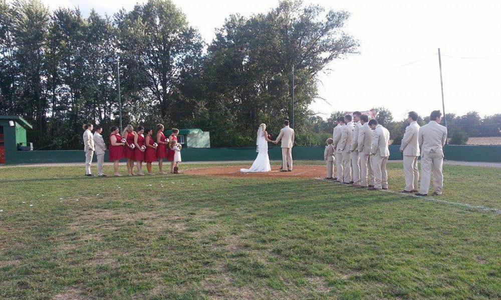 A photo of Ross and Nikki Westdorp's wedding at Parker Field. The couple stands at homeplate. The bridesmaids stand along the first base line. The groomsmen stand on the third base line.