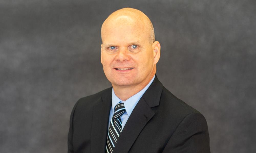 A headshot of Alex Cline. He's wearing a black suit while a light blue shirt and a tie with white and light blue stripes.