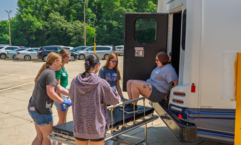 Students at the Pathways to Healthcare Camp participate in a simulated mass casualty incident, working together to safely transfer a mock patient from an ambulance to a stretcher in a parking lot.