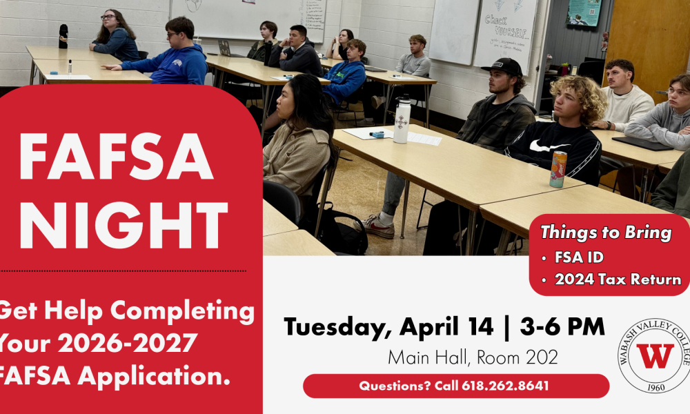A classroom of students seated at individual desks arranged in a U‑shaped layout, all facing toward the front of the room. A whiteboard is visible on the wall with drawings and notes, and classroom items such as notebooks, laptops, water bottles, and small supplies are on the desks. The room has white brick walls and a tan floor.