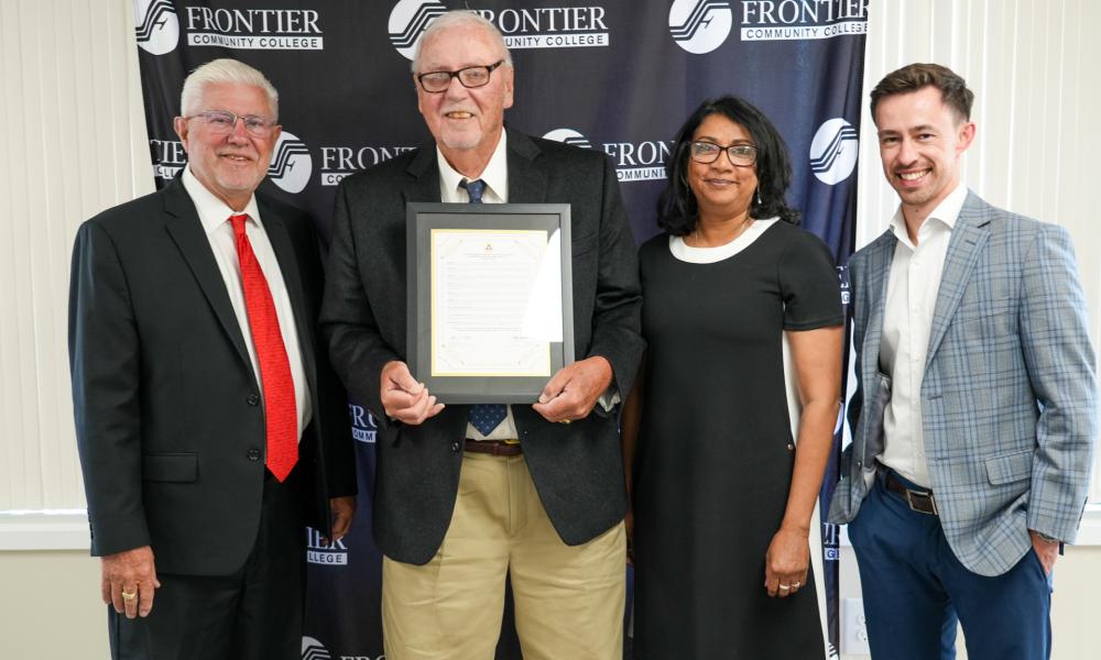 IECC Board Chairman Gary Carter, Jerry Cox holding a framed proclamation in his honor, FCC President Dr. Sharmila Kakac, and FCC Foundation President Luke Harl stand together in front of a Frontier Community College backdrop.