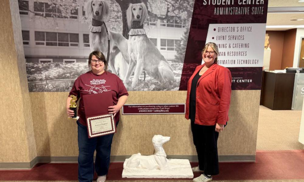 Jessica Wagner (left) holds a certificate and award while standing beside Amie Mayhall (right). They pose in a campus hallway, celebrating Wagner’s accomplishments at the SIU Accounting Competition, with a dog statue and display signage behind them.