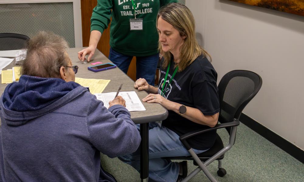 An IECC staff member sits with an older adult at a table, reviewing paperwork together and offering guidance, while another staff member stands nearby assisting.
