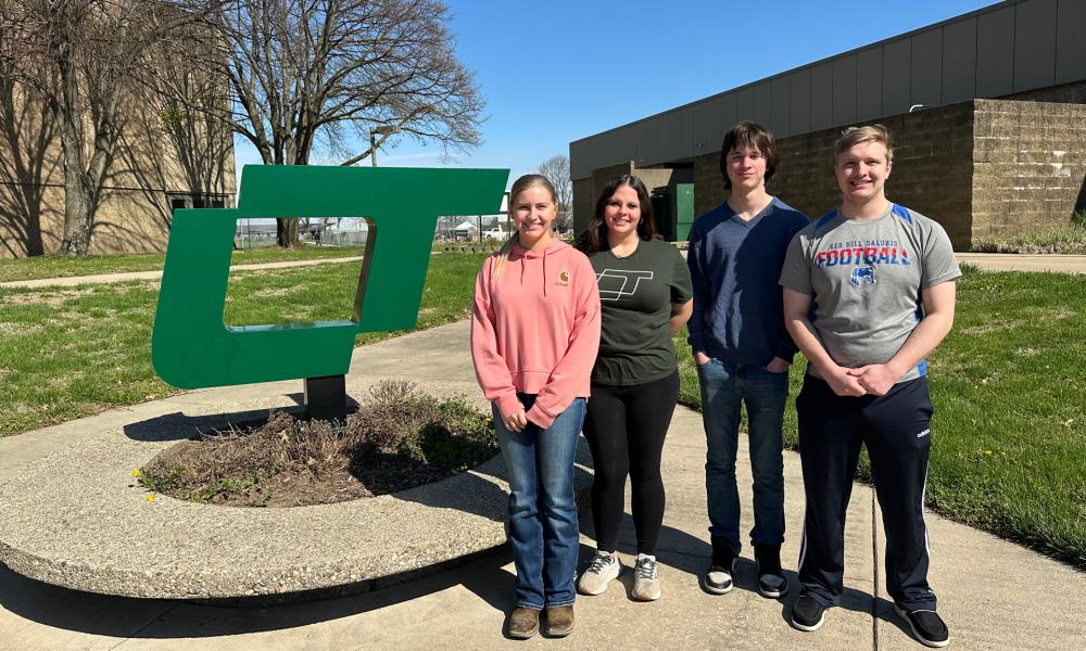 Four Lincoln Trail College students—Hannah Potts, Natalie Ramsey, Korbin Alger, and Dalton Spahn—stand outdoors near a large green LTC logo on a sunny day, smiling for a group photo.