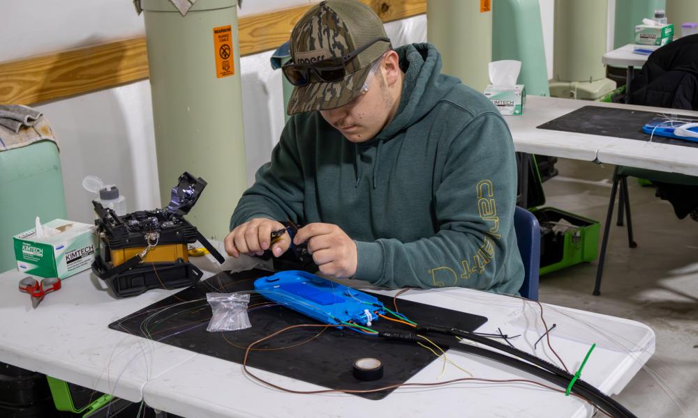 A Broadband Telecom student sits at a workstation carefully splicing fiber optic cables, using specialized tools and equipment spread across a table during hands-on training.