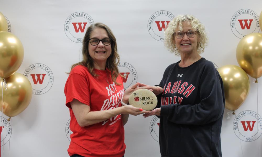 Director of Learning Commons and her assistant stand holding the NILRC Rock in front of a WVC backdrop with gold balloons flanked on either side.