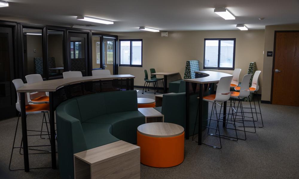 Collaborative workspace at The 618, featuring green lounge seating, orange ottomans, high-top tables and white chairs with orange cushions. Glass-walled meeting rooms line one side of the space.