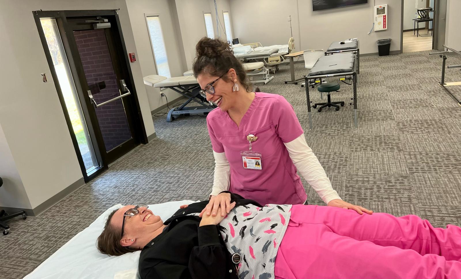 PTA Students in lab. One student laying down the other hovering over the one laying down. Both smiling