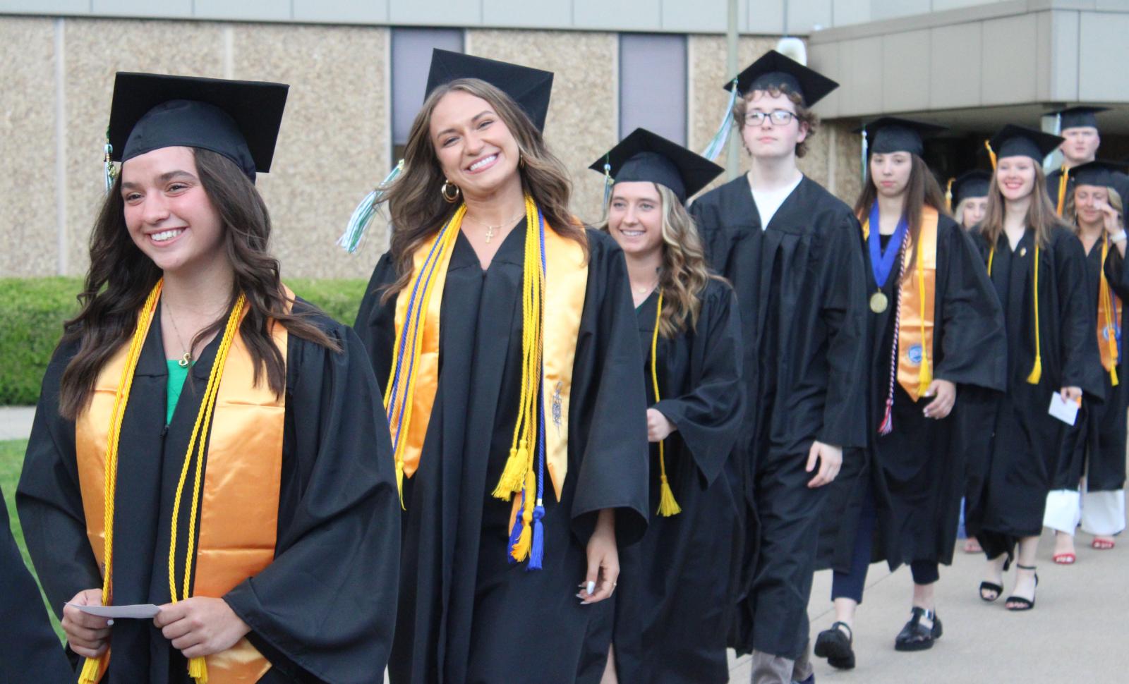 A group of graduates in black caps and gowns walk in a line outside a building, smiling and celebrating. Some students wear gold stoles and honor cords, indicating academic achievements.