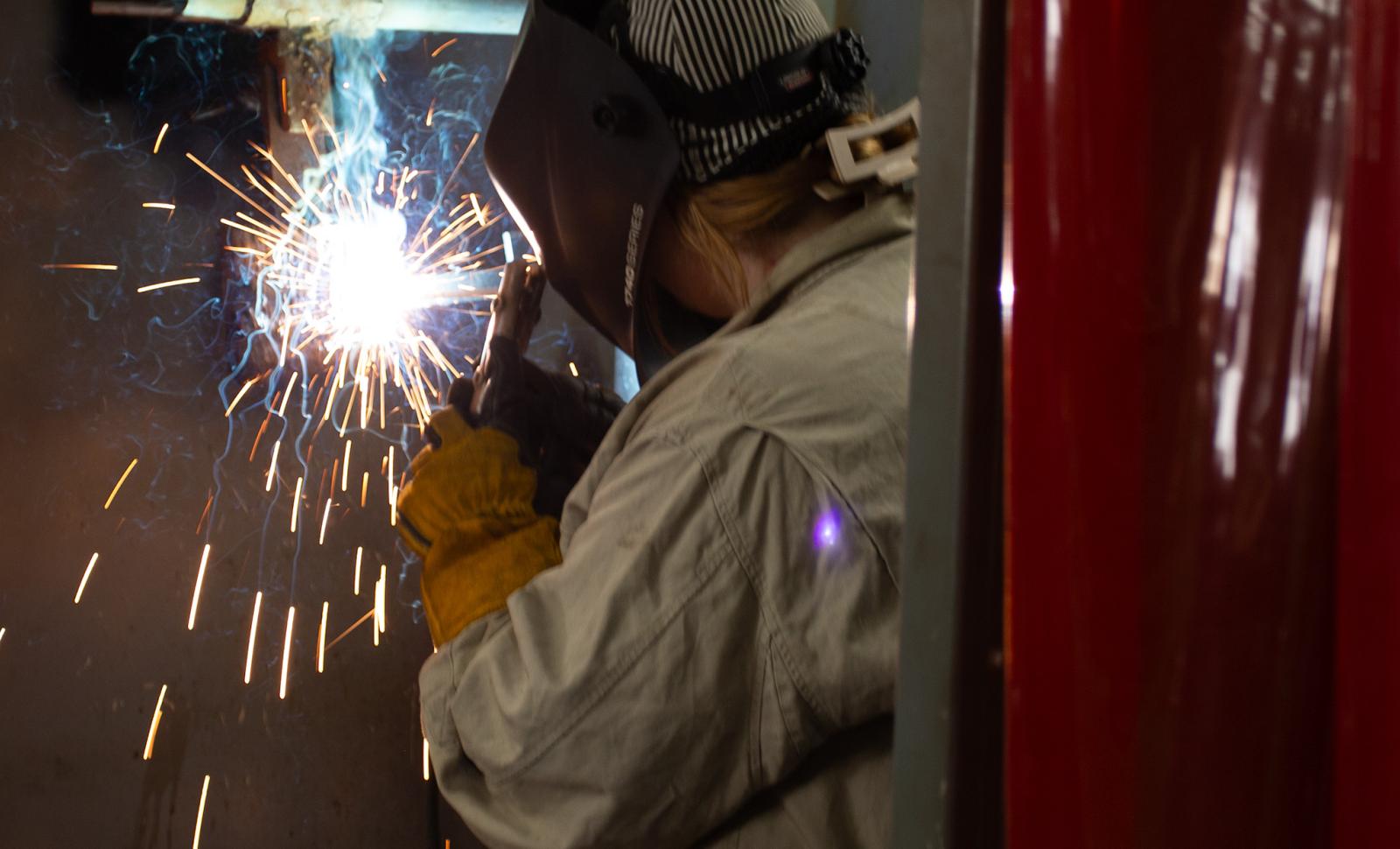 A welding student wearing protective gear, including a welding helmet and gloves, works inside a welding booth. Bright sparks fly from the welding torch as the student welds a metal pipe, with red welding curtains visible on both sides.