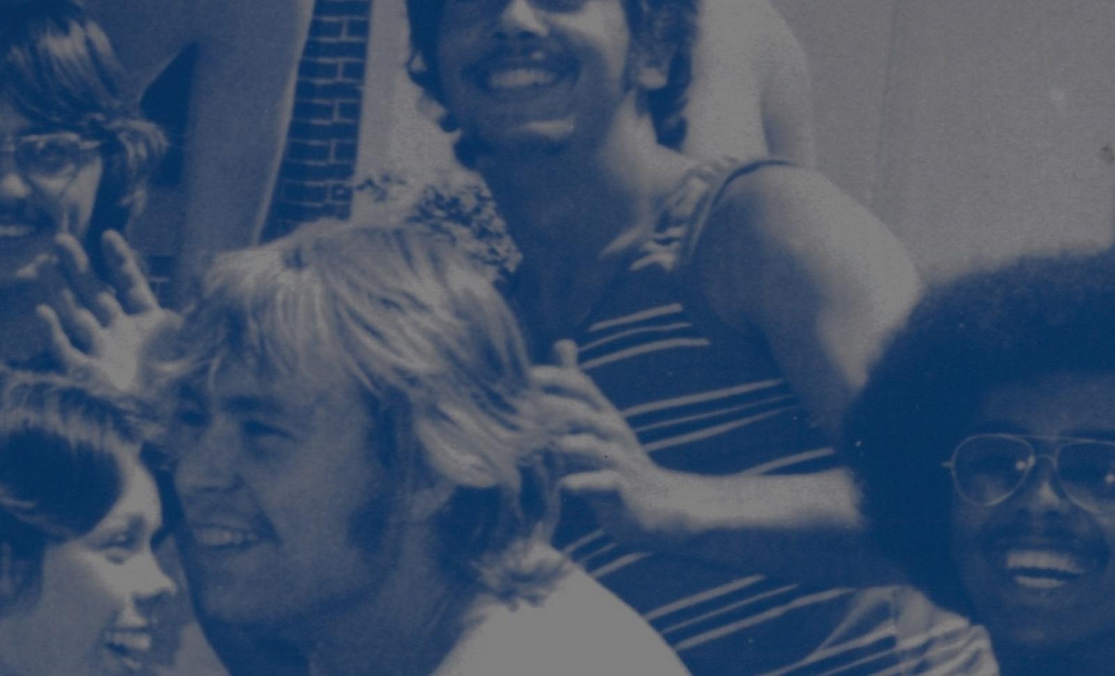 Vintage blue-tinted photo of a group of smiling college students gathered outside a brick building, laughing and posing closely together.