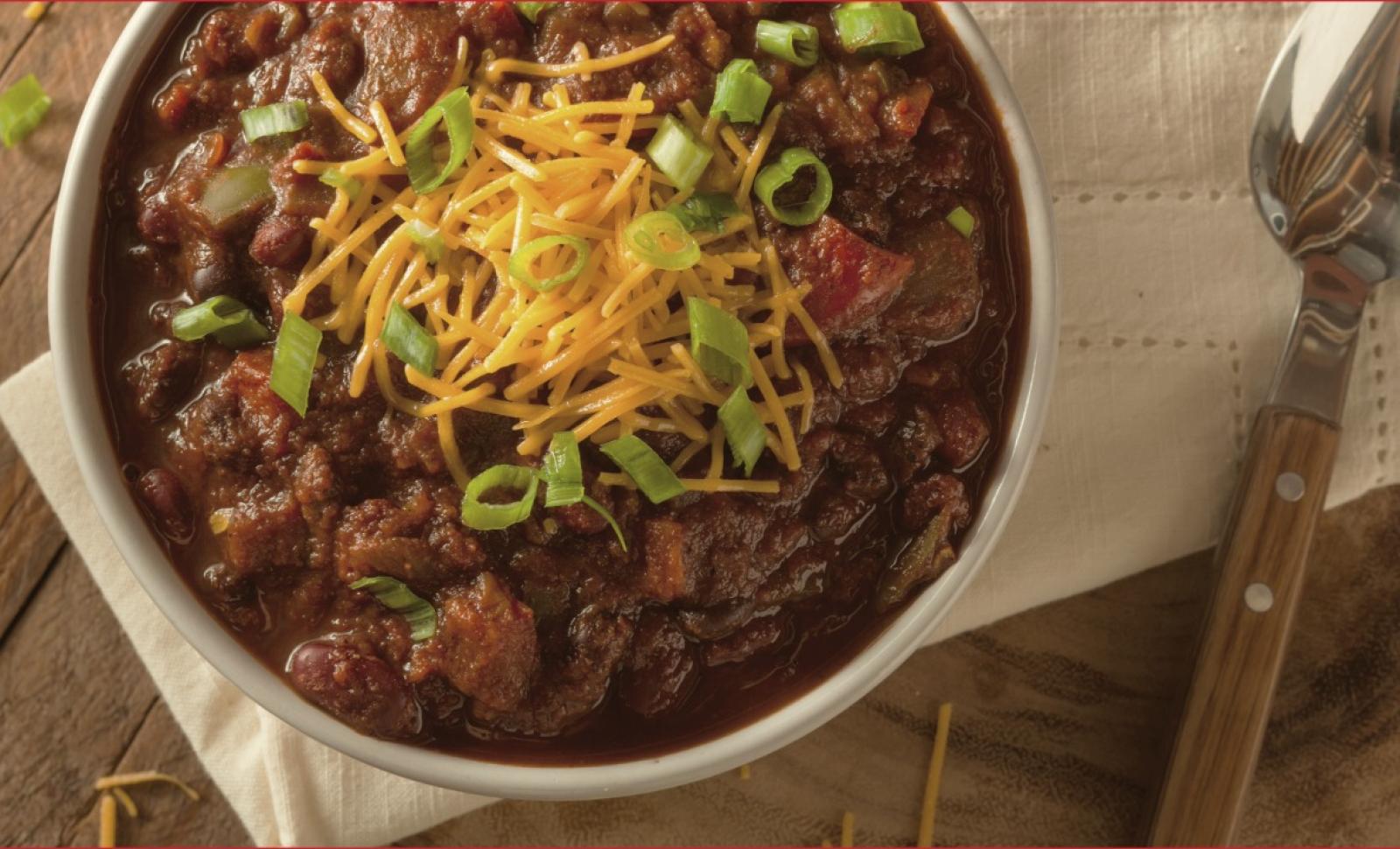 Image of a bowl of chili with cheese and green onions and a spoon to its right