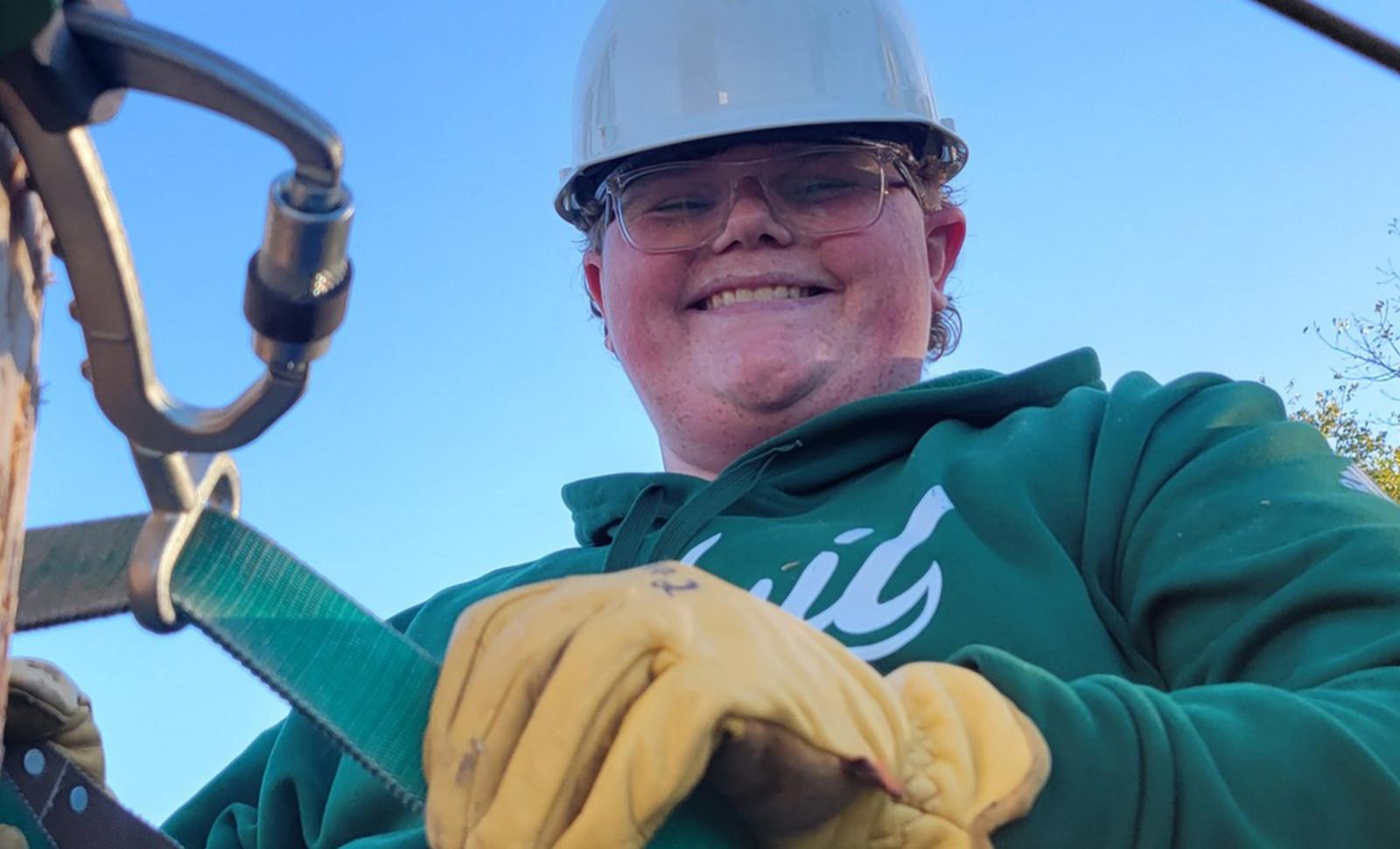 Student wearing a hard hat, safety glasses, and work gloves smiles while fastening a safety strap to a wooden utility pole outdoors. The student is dressed in a green sweatshirt and appears to be participating in hands-on technical or utility training.