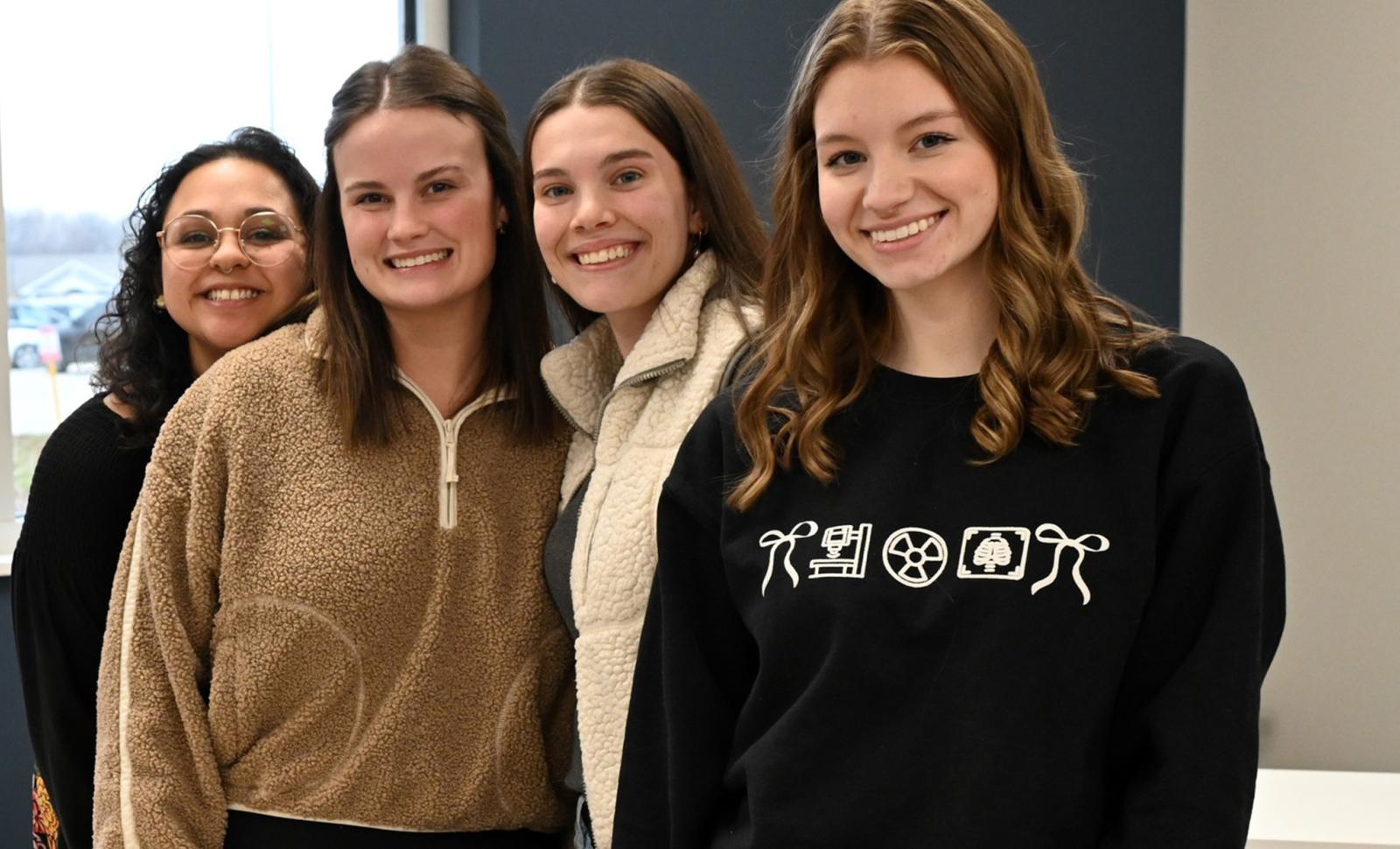 Four female students stand side by side indoors, smiling at the camera in a classroom setting. A window behind them shows a parking lot outside. A medical training mannequin torso is visible on a table in the foreground, suggesting a health sciences or lab environment.