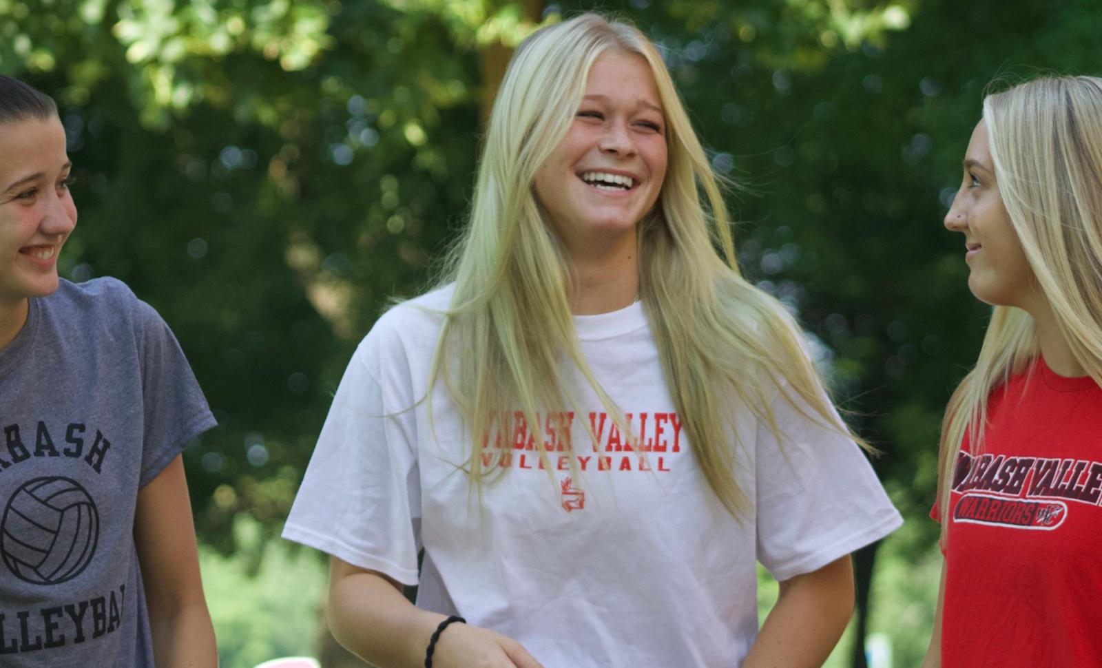 Three female students stand outdoors in a wooded campus setting, smiling and laughing together. They are wearing Wabash Valley College T-shirts, including volleyball and athletics apparel, as they talk and enjoy time together.