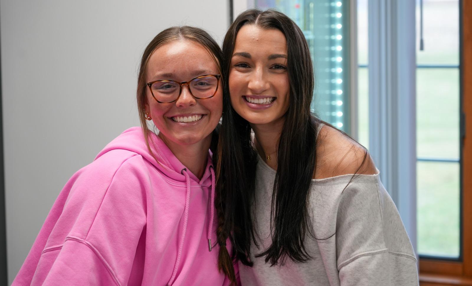 Two young women smiling and posing closely together indoors; one is wearing glasses and a pink hoodie, and the other has long dark hair and is wearing a light-colored off-shoulder top, with a softly lit window in the background.