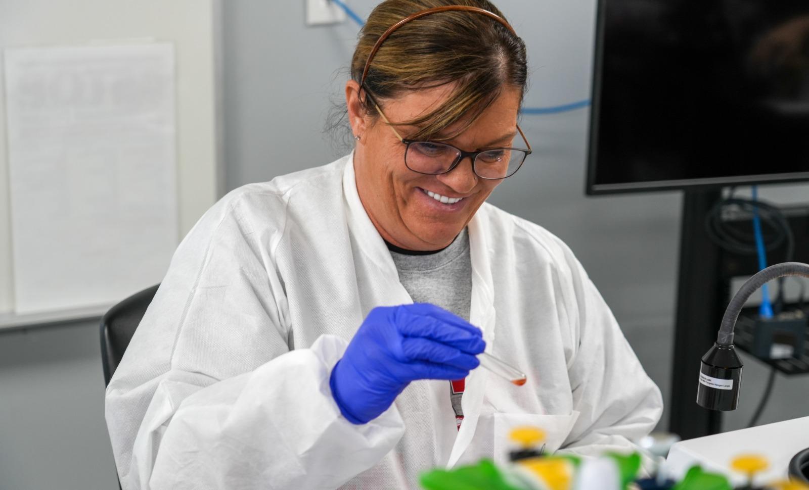A woman wearing glasses, a lab coat, and blue gloves smiles while using a dropper to handle a small liquid sample in a laboratory setting.