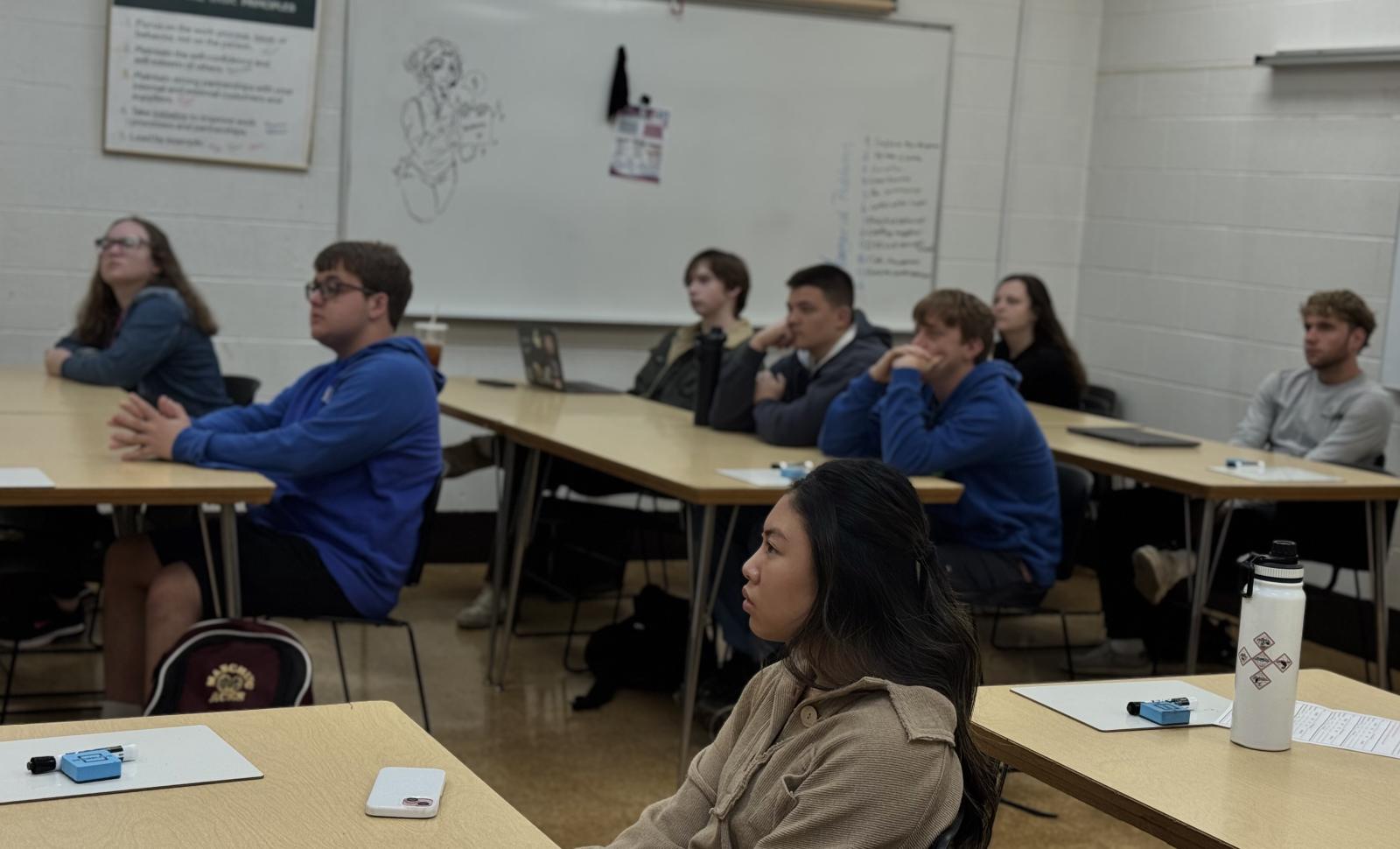 A classroom of students seated at individual desks arranged in a U‑shaped layout, all facing toward the front of the room. A whiteboard is visible on the wall with drawings and notes, and classroom items such as notebooks, laptops, water bottles, and small supplies are on the desks. The room has white brick walls and a tan floor.