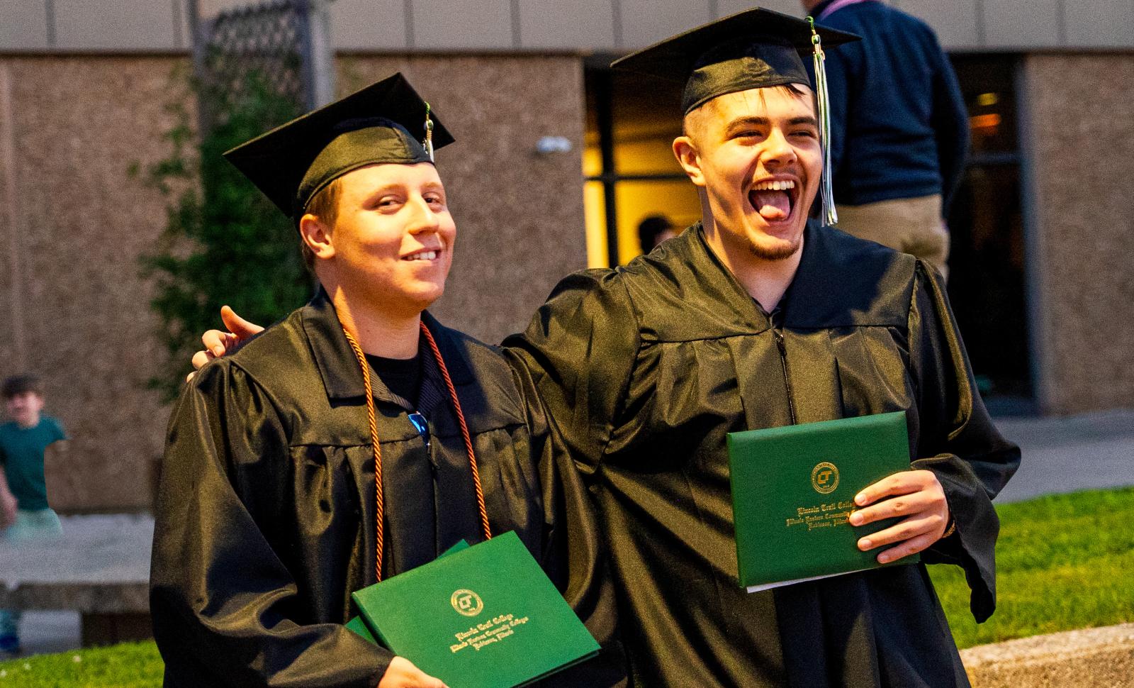 Two graduates in caps and gowns stand arm in arm, smiling and celebrating while holding green diploma covers outside a building.