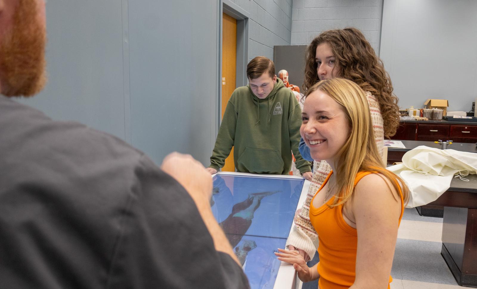 Students gather around an interactive anatomy table in a classroom as an instructor gestures, with a model of the human body and lab materials visible in the background.