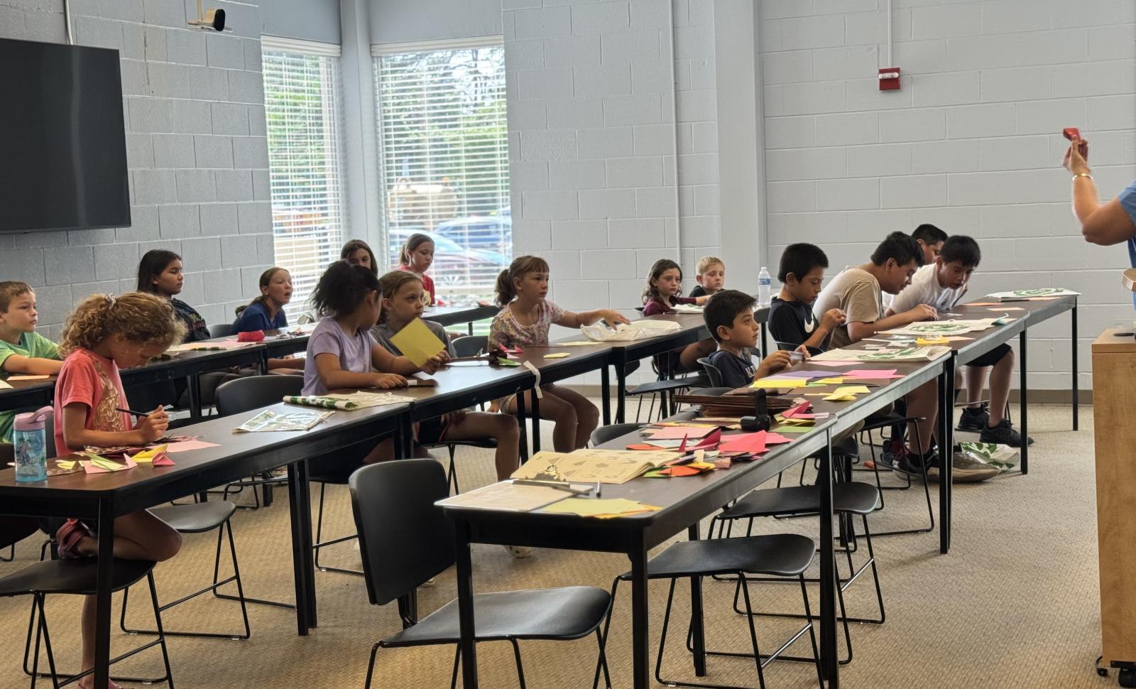 Classroom scene with students seated at tables working on hands-on activities while an instructor stands at a podium leading the lesson. The room includes long tables, chairs, craft materials, windows, and a wall-mounted screen.