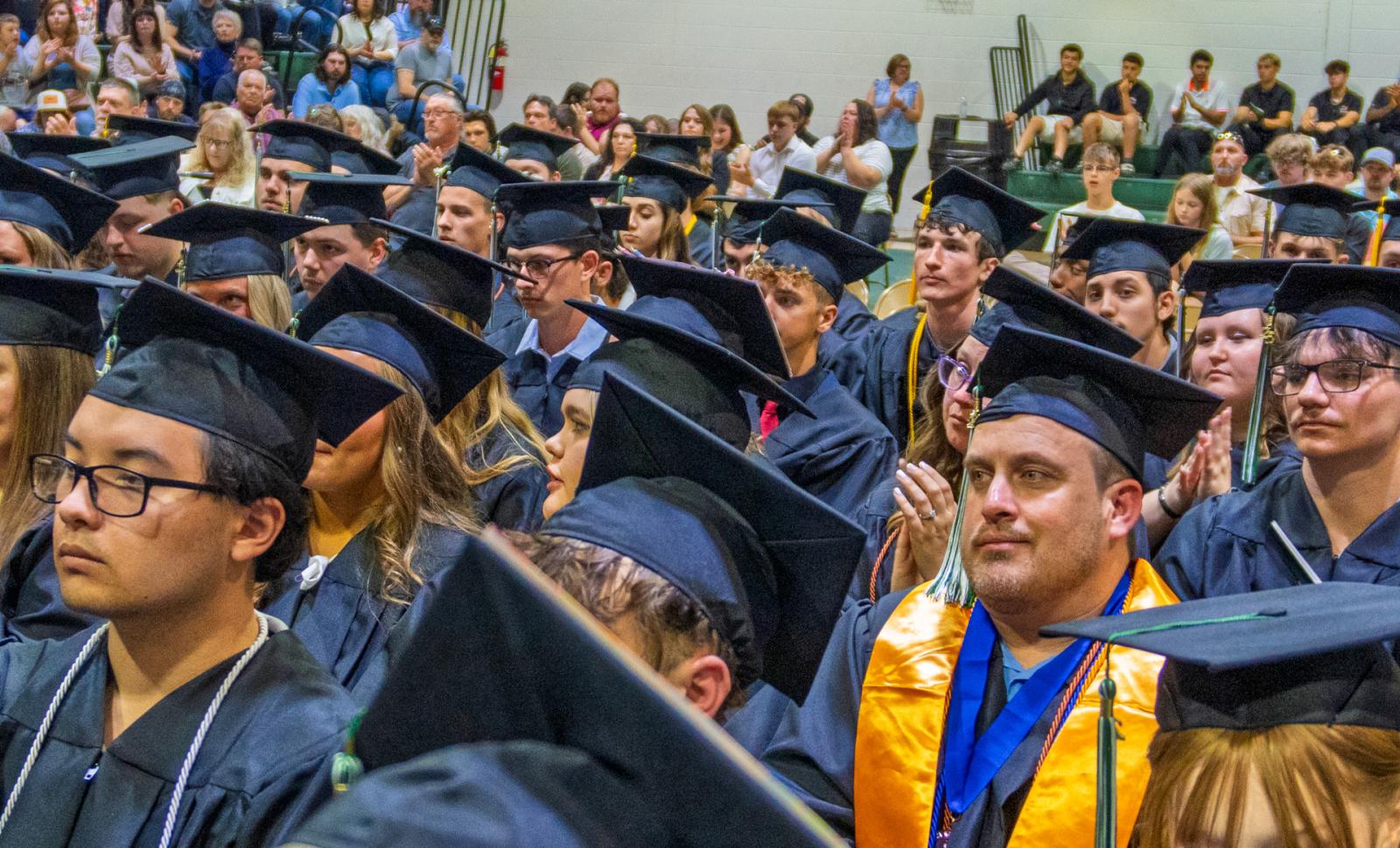 Lincoln Trail College graduates sit in caps and gowns during a commencement ceremony, listening attentively as audience members in the background applaud.