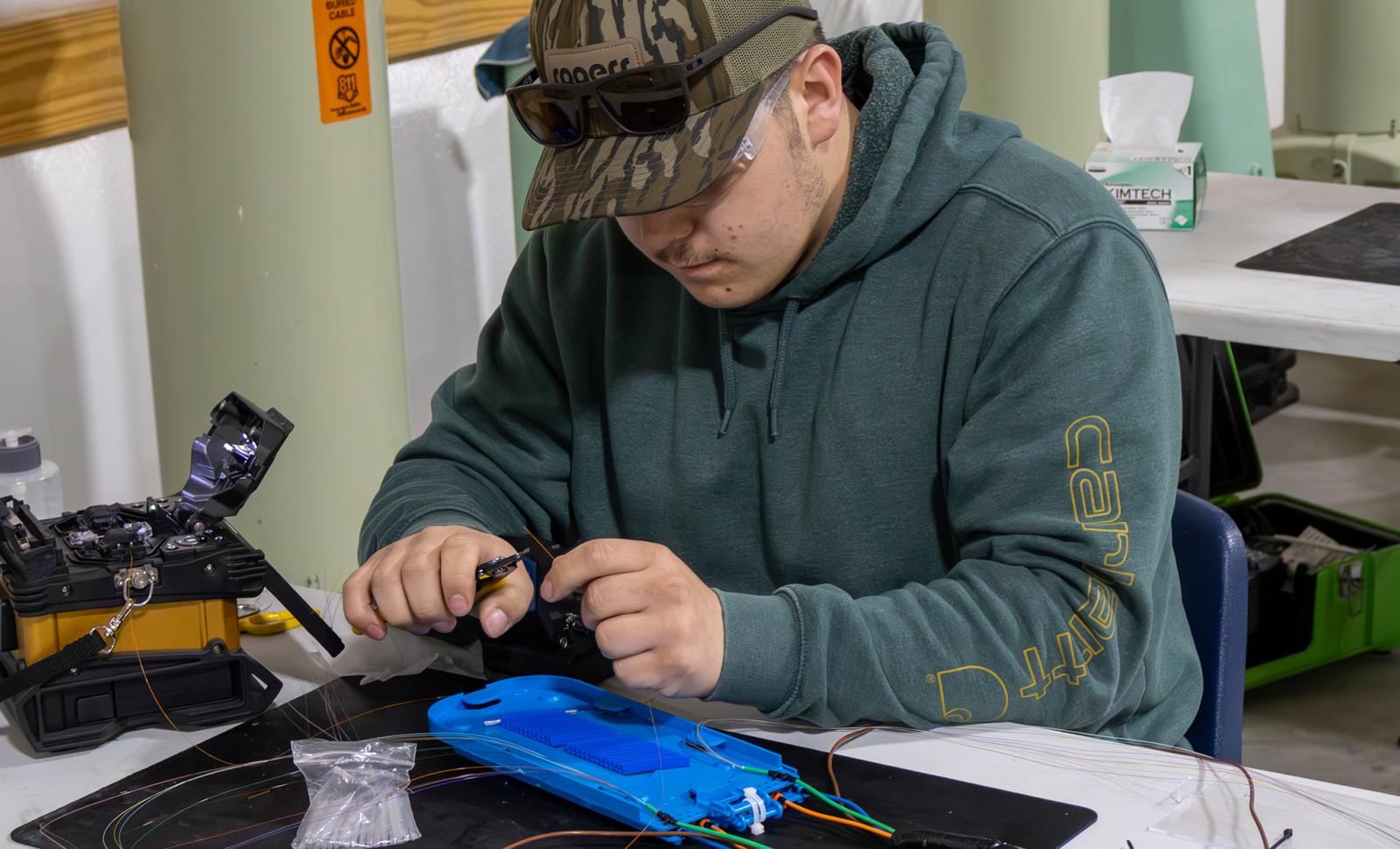 A Broadband Telecom student sits at a workstation carefully splicing fiber optic cables, using specialized tools and equipment spread across a table during hands-on training.