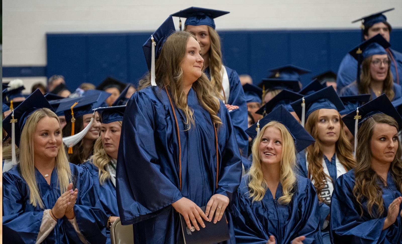Student in cap and gown standing among the other seated graduates