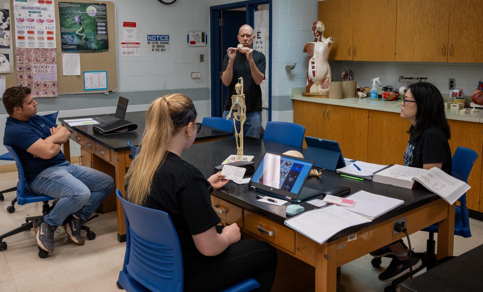 Anatomy Professor Chris Mathews holds up a bone during a lecture. Students are sitting at tables observing.
