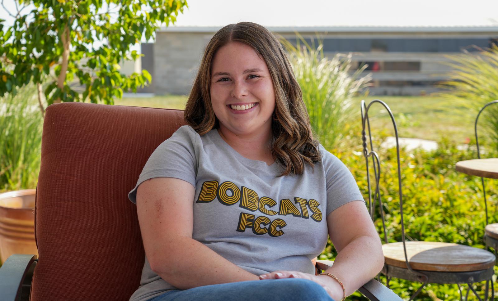 A smiling woman wearing a gray FCC Bobcats T-shirt sits in an outdoor chair with greenery and a campus building behind her.