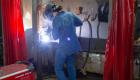 A welding student wearing a protective helmet and denim clothing is actively welding a metal piece in a workshop. Bright sparks and a welding arc are visible. Red safety curtains surround the work area, and various welding tools and equipment are present.