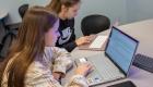 Two students in a business class at Illinois Eastern Community Colleges sit at a table working on laptops. One student types on a silver laptop while the other follows along on a rose-gold laptop, both focused on an online assignment displayed on their screens in a classroom setting.