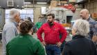 A group of adults stands in a welding lab during an open house, talking with welding instructor Maverick Fisher wearing a red shirt. Community members listen and ask questions while surrounded by shop equipment, worktables, and ventilation systems, highlighting a hands-on learning environment.